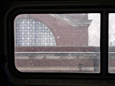 [The image is nearly completely outlined by the framing of the window. Outside the window is a red brick building with white x-framed windows. White chunks of snow are flying between the window and the building.]